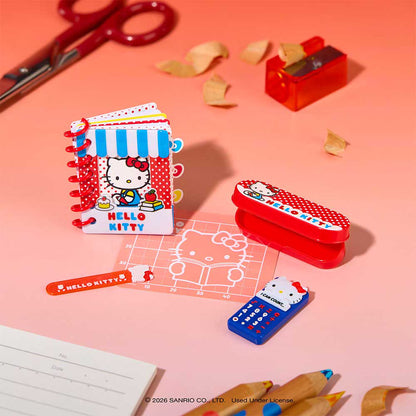 Hello Kitty themed stationery set on a pink background, including a small notebook, red eraser, blue calculator, clear grid ruler, and pencil sharpener, with scissors and paper scraps nearby.