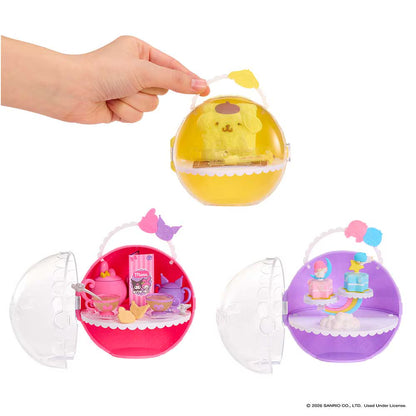 Three round translucent toy cases shown on a white background, with a human hand holding the top yellow case open. The cases contain miniature dollhouse-style accessories and are shown in yellow, pink, and purple colorways.