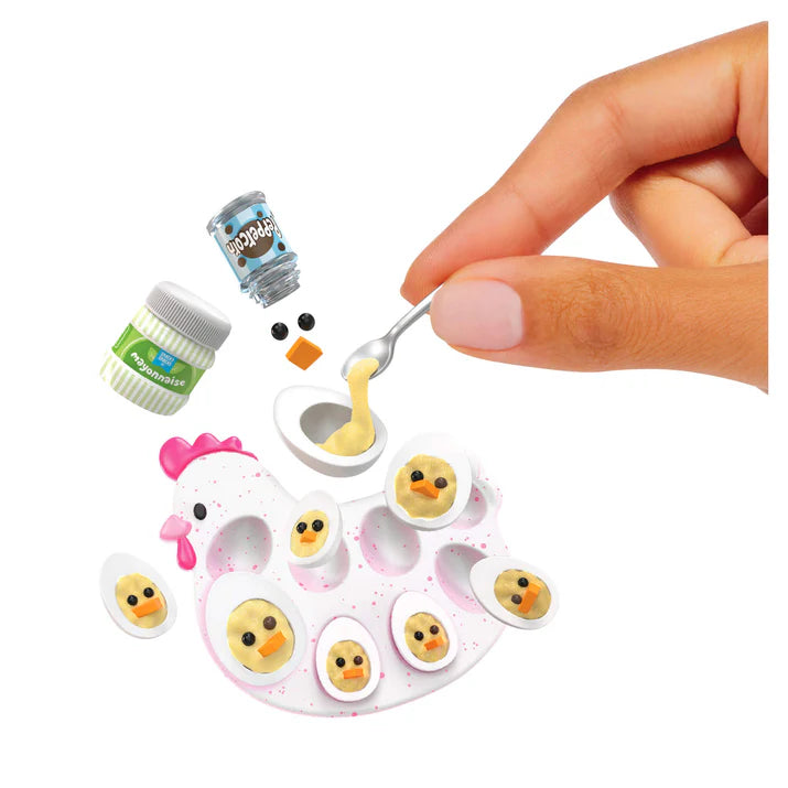 A hand using a silver spoon to fill a miniature egg white with yellow "yolk" resin to create the Chick Deviled Eggs, alongside a tiny jar of mayonnaise and peppercorns.