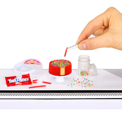 A hand using white tweezers to place a miniature red Twizzler stick onto a tiny cake base topped with sprinkles and surrounded by other Twizzlers ingredients.