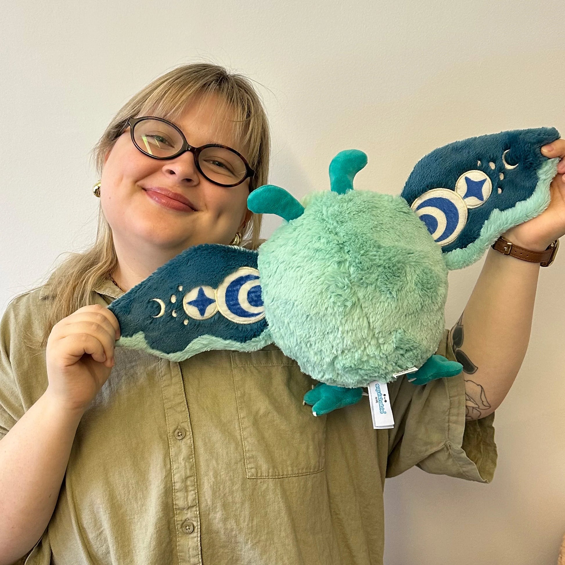 A person wearing glasses and a khaki button-up shirt holds a plush moth toy. The toy has a mint green fuzzy body with dark teal wings featuring embroidered white moon and star patterns.