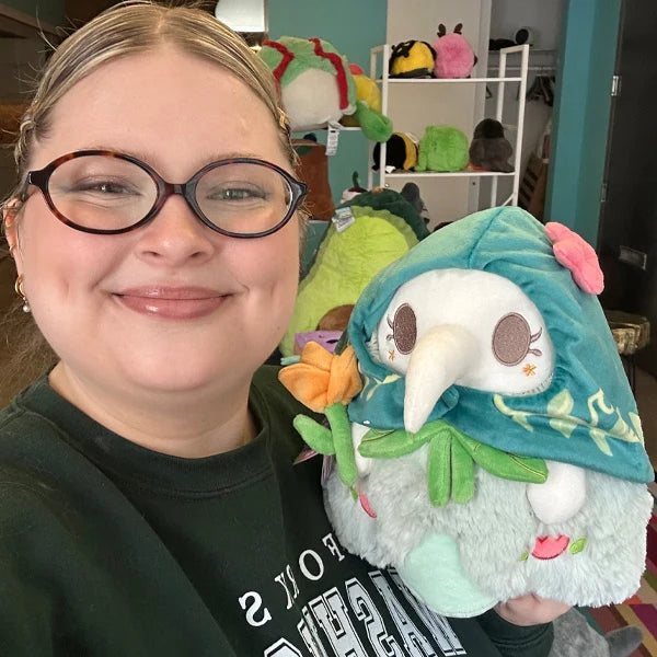 Person holding Mini Squishable Botanical Plague Nurse plush with teal vine-embroidered hood, white beak, light green dress, and holding an orange flower in a room with shelves of more plush toys.