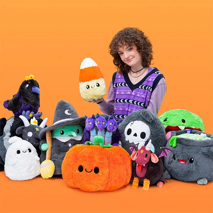 A person stands against a solid orange background holding a cute plush candy corn toy. In front of them is a large collection of various colorful Halloween-themed plush toys, including a pumpkin, a witch, a raven, a ghost, and a cauldron.