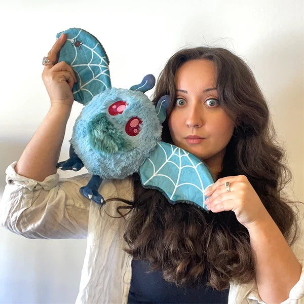 A woman with long brown hair holds a Mini Squishable Haunted Mothman plush featuring light blue fur, glowing red eyes, and dark tattered wings.