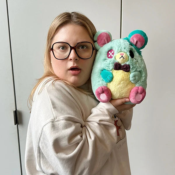 A person wearing black-rimmed glasses and a beige sweatshirt holds a pastel plush toy mouse. The plush has mint green, yellow, pink, and teal colors with embroidered facial details.