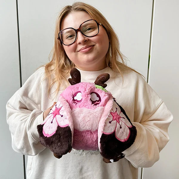 A woman with blonde hair and glasses holds a pink plush owl mitten with dark brown accents and cherry blossom details, wearing a cream sweatshirt against a light gray paneled background.