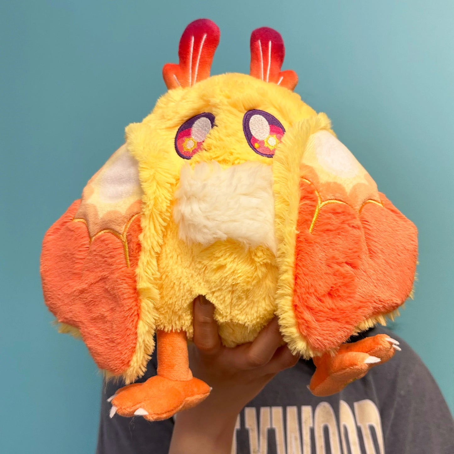 Plush toy rMini Squishable Solar Mothman plush featuring bright yellow fur, orange wings, and red gradient antennae. with large eyes held by a person against a blue background