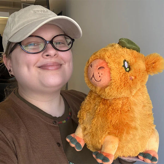 Person wearing a cap and glasses holding an orange plush toy with a green leaf.