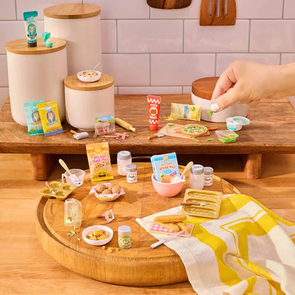 Miniature food items and kitchen accessories on a wooden table with a hand interacting with them. Miniverse Make It Mini From Scratch Series 2