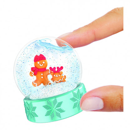 A small clear snow globe with glitter-filled sphere, featuring gingerbread characters inside, held by two fingers against a plain white background.