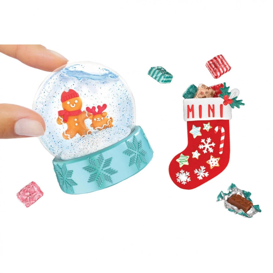 A hand holds a clear snow globe ornament with a light blue base featuring a gingerbread man and reindeer design. To the right are several small wrapped candy pieces and a red Christmas stocking ornament labeled "MINI" with white snowflake and candy cane details, all on a plain white background.