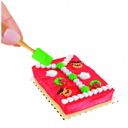 A miniature toy cake with bright red icing, decorated with white piped borders, green accents, and small character cookies, being lifted by a hand holding a small spatula on a white background.