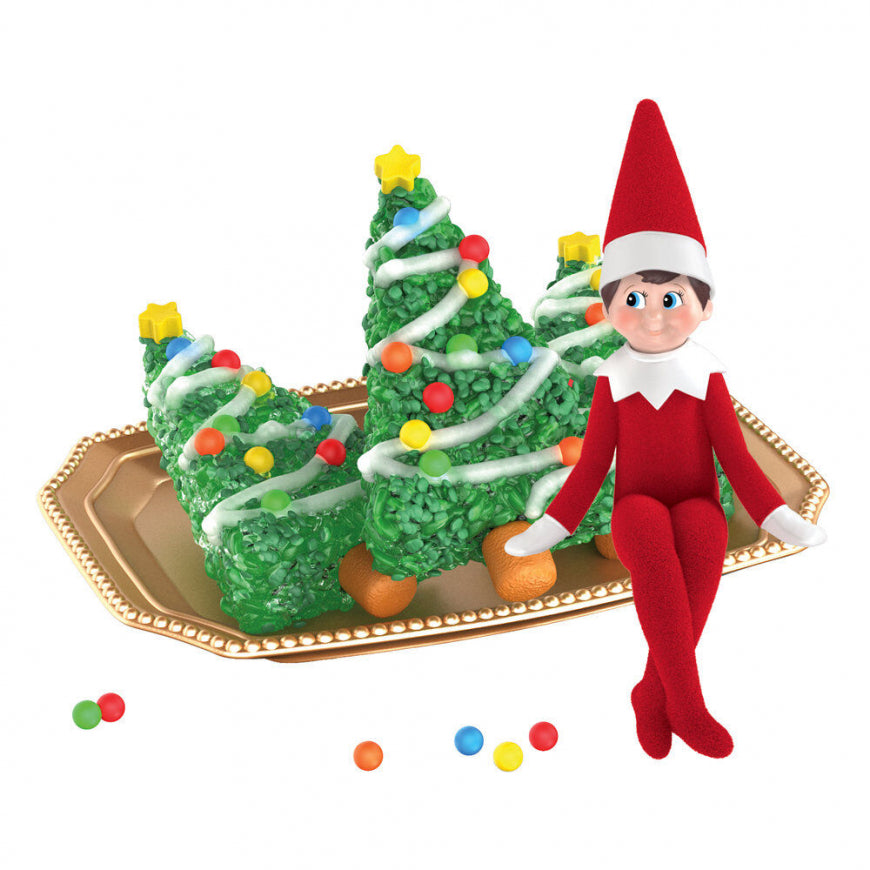 A festive holiday scene featuring three decorated green rice krisp trees on a gold tray, alongside a small red elf doll sitting on the tray. Loose candy pieces are scattered on the white background.