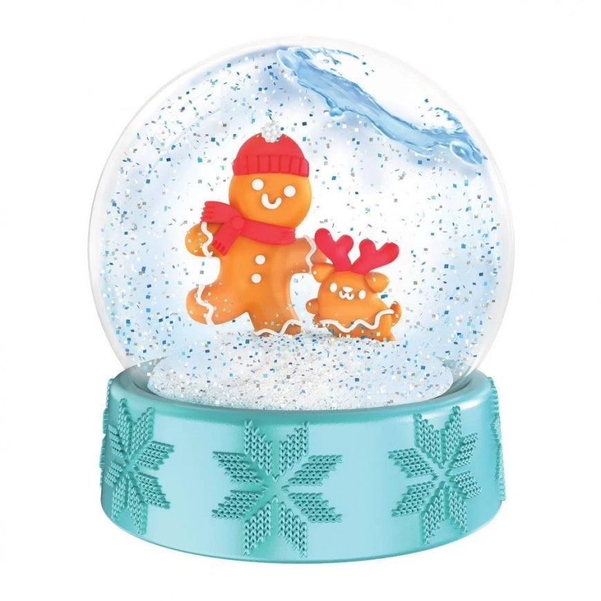 A festive gingerbread snow globe featuring a large clear sphere filled with white glitter and blue decorative swirls, containing two orange gingerbread figures—a larger one wearing a red hat and scarf and a smaller one with red antlers. The base is light aqua with embossed snowflakes.