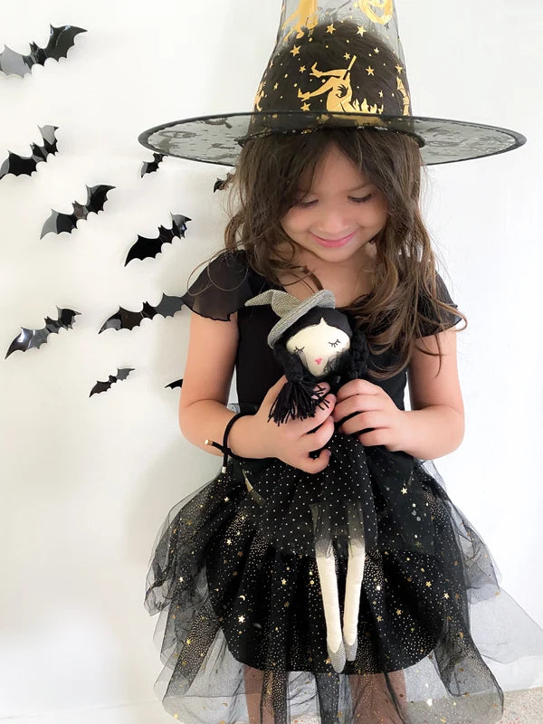 A young girl wearing a black and gold witch costume with a large pointed witch hat, holding a small black-and-white witch doll against a white wall decorated with hanging black bats.