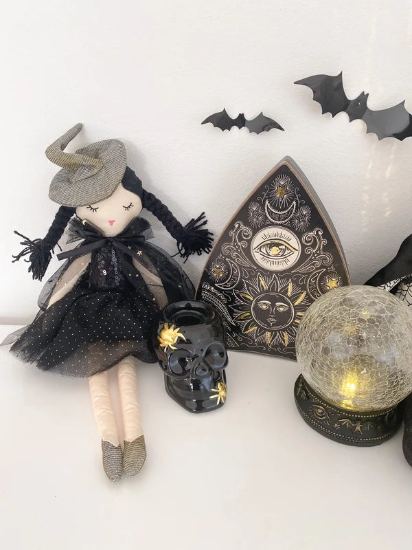A styled Halloween display featuring a witch doll in a black dress and grey hat next to a black crystal ball lamp with a warm light, a black skull candle holder, and a triangular black and gold celestial board, all set against a white background with black bat cutouts.