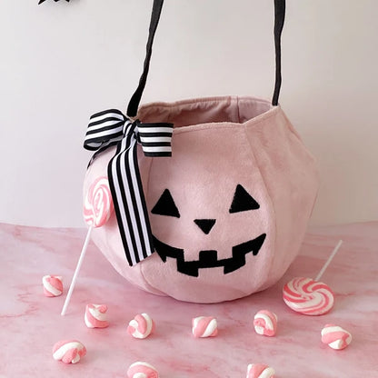 A pink plush pumpkin-shaped candy bucket with a black embroidered jack-o'-lantern face, black handles, and a black-and-white striped ribbon bow. Pink and white lollipops and candy pieces are arranged around it on a pink surface against a light background.