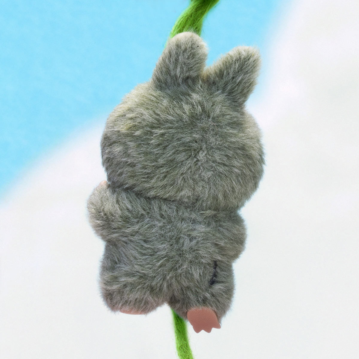 Plush toy shaped like a gray creature with pink feet on a green stem against a light blue and white background.