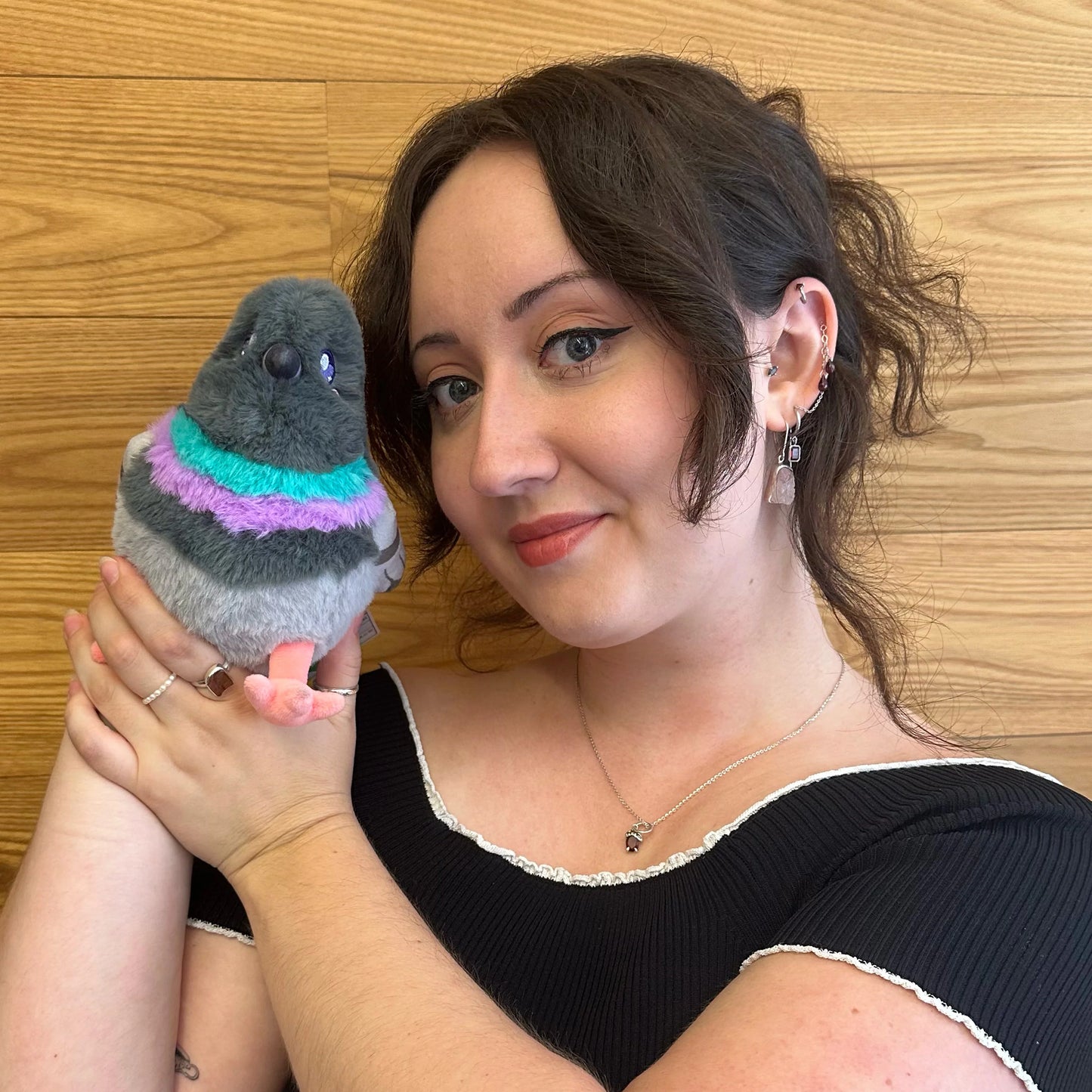 A woman with dark hair holding a small plush stuffed pigeon with a gray body and a colorful turquoise and purple striped neck, posed against a light wood-paneled wall.