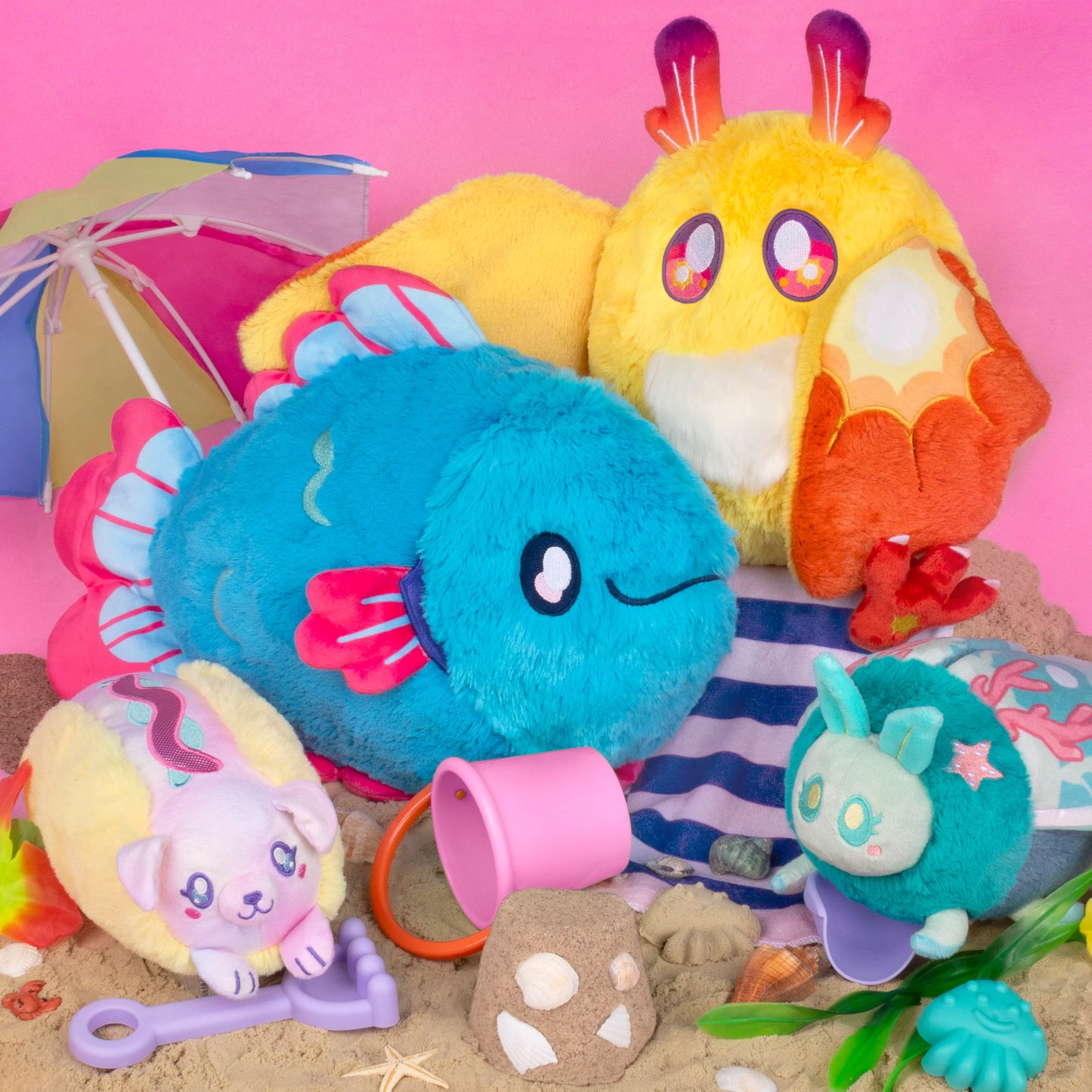 A collection of colorful plush toys arranged on sand against a pink background.
