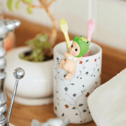 Cute figurine of a baby with a green helmet and toothbrush in a floral-patterned cup on a wooden surface.