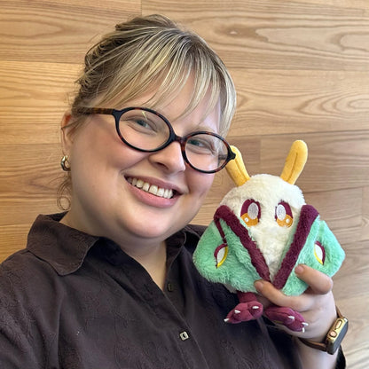 A smiling woman with glasses holds a Squishable Alter Egos Series 21 Luna Mothman plush with cream fur, yellow antennae, burgundy feet, and mint green wings, posed in front of a light wood wall.
