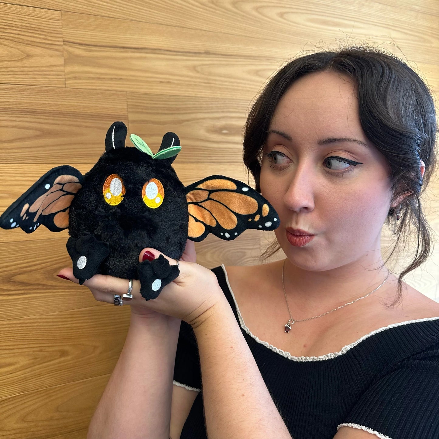 A woman holding aSquishable Alter Egos Series 21 Monarch Mothman plush with black fur, orange glowing eyes, a leaf accent, and monarch butterfly wings posed in front of a light wood-paneled wall.