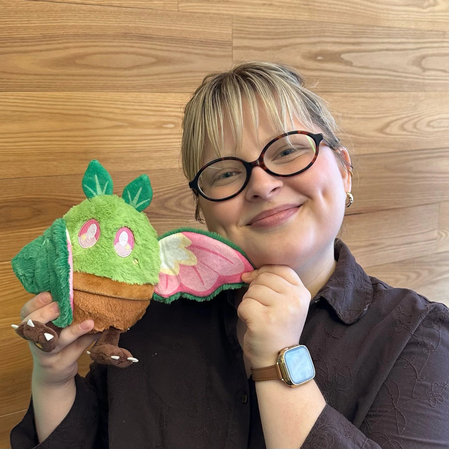 A person with short blonde hair and black-rimmed glasses holdsSquishable Alter Egos Series 21 Plant Mothman plush sitting in a plush brown flowerpot with bright green fur, leafy antennae, and leafy wings against a light wood-paneled background.
