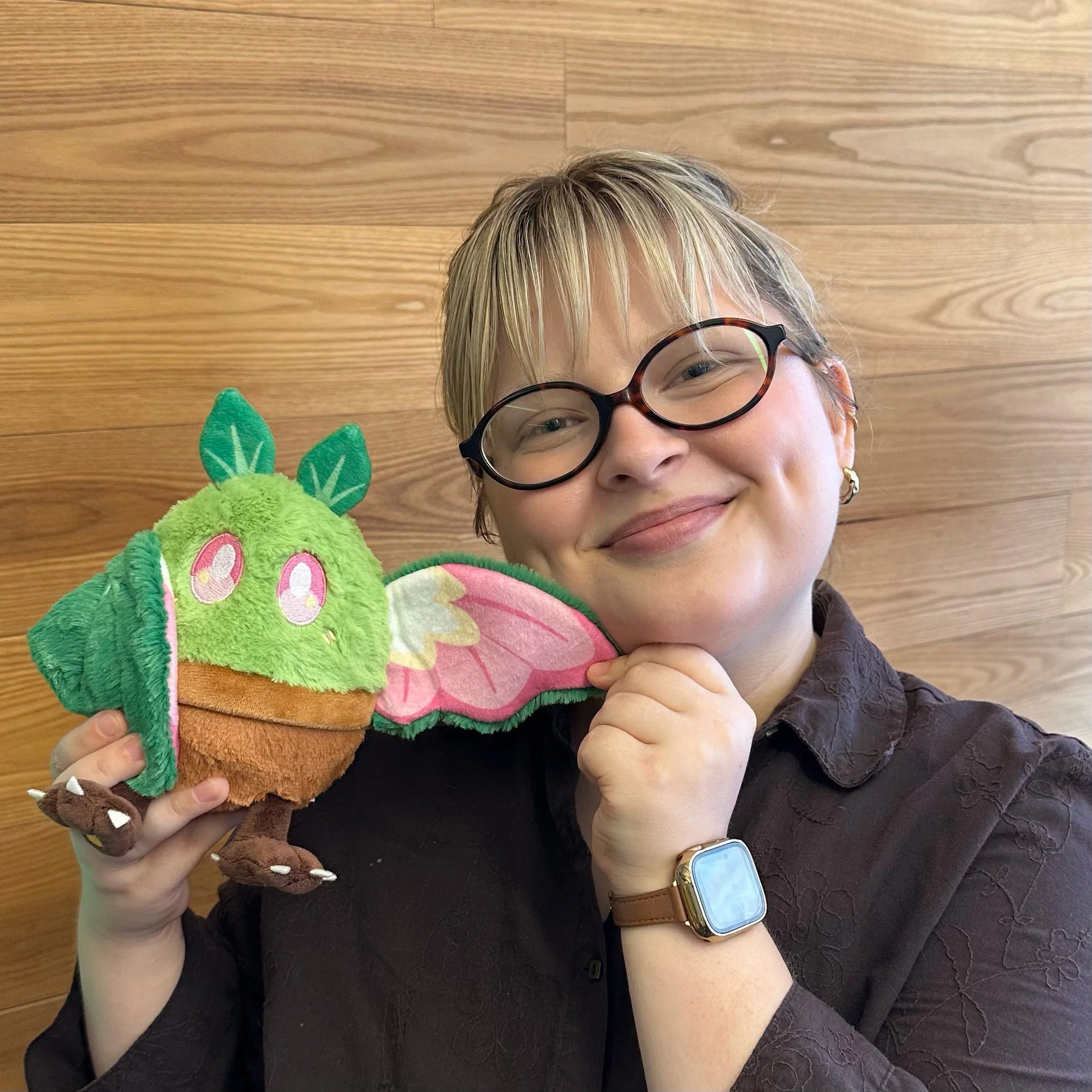 A person with short blonde hair and black-rimmed glasses holdsSquishable Alter Egos Series 21 Plant Mothman plush sitting in a plush brown flowerpot with bright green fur, leafy antennae, and leafy wings against a light wood-paneled background.