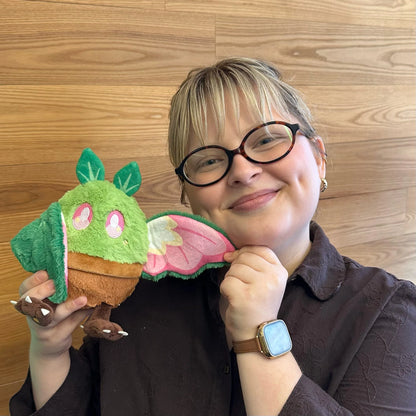 A person with short blonde hair and black-rimmed glasses holdsSquishable Alter Egos Series 21 Plant Mothman plush sitting in a plush brown flowerpot with bright green fur, leafy antennae, and leafy wings against a light wood-paneled background.