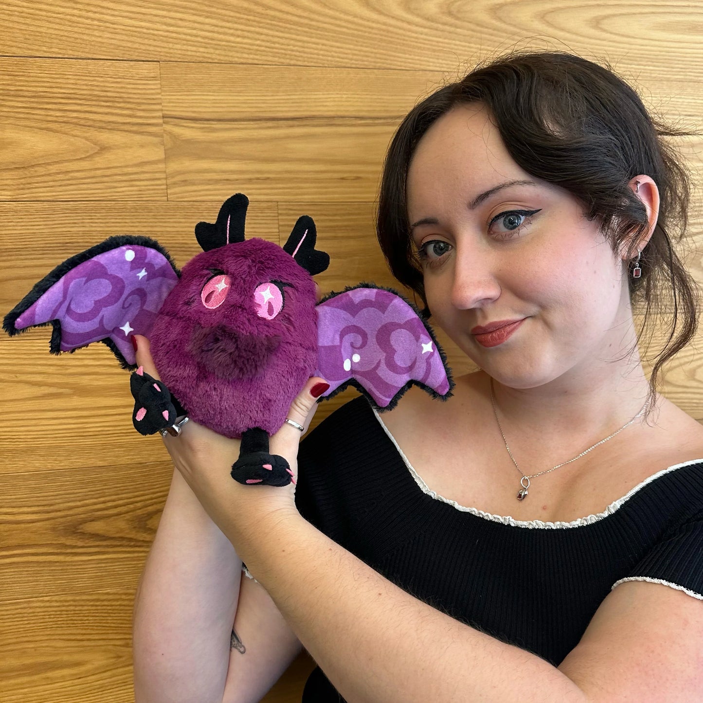 A woman holding a Squishable Alter Egos Series 21 Poison Mothman plush featuring deep magenta fur, glowing pink eyes, and black wings with pink embroidery posed against a light wood-paneled wall.