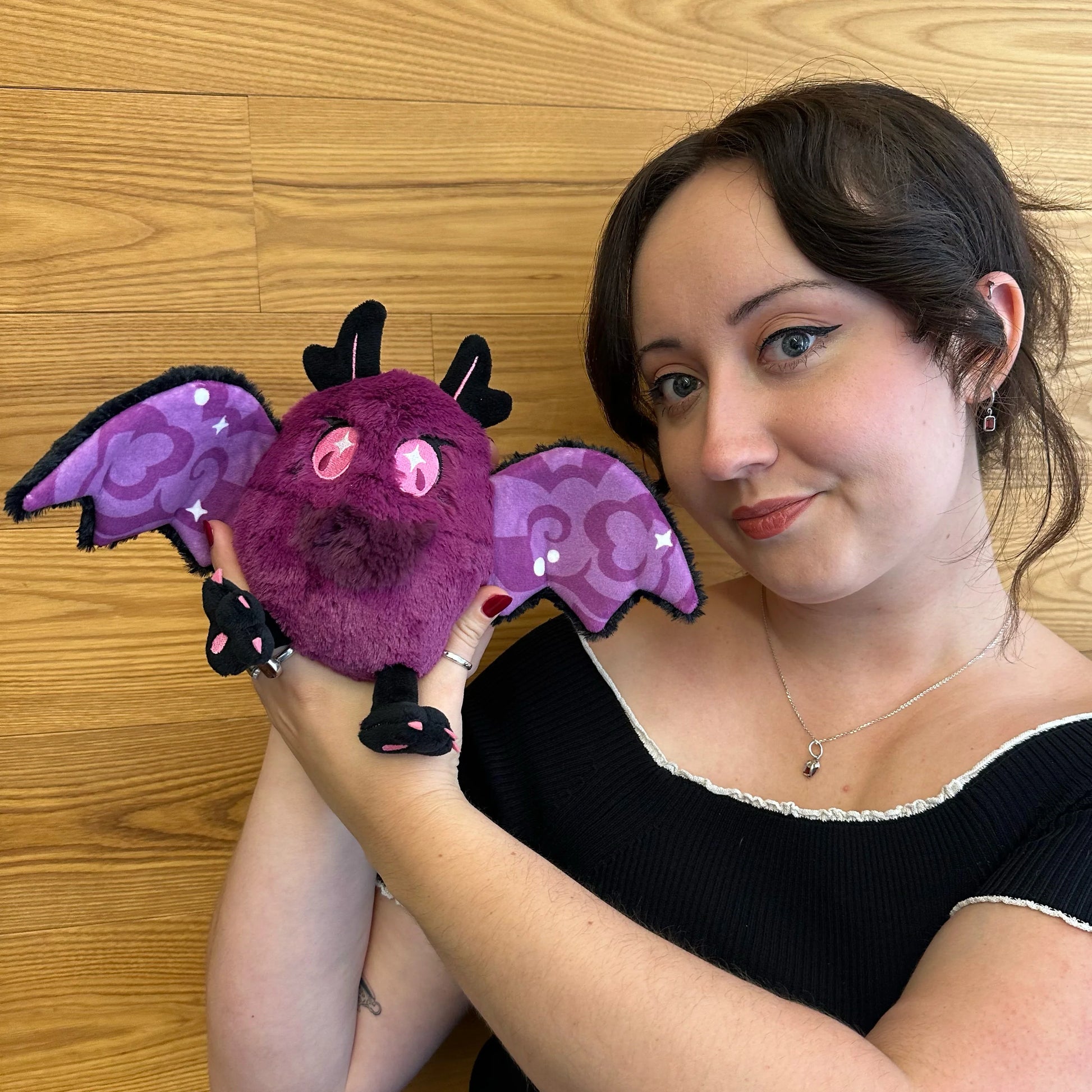 A woman holding a Squishable Alter Egos Series 21 Poison Mothman plush featuring deep magenta fur, glowing pink eyes, and black wings with pink embroidery posed against a light wood-paneled wall.