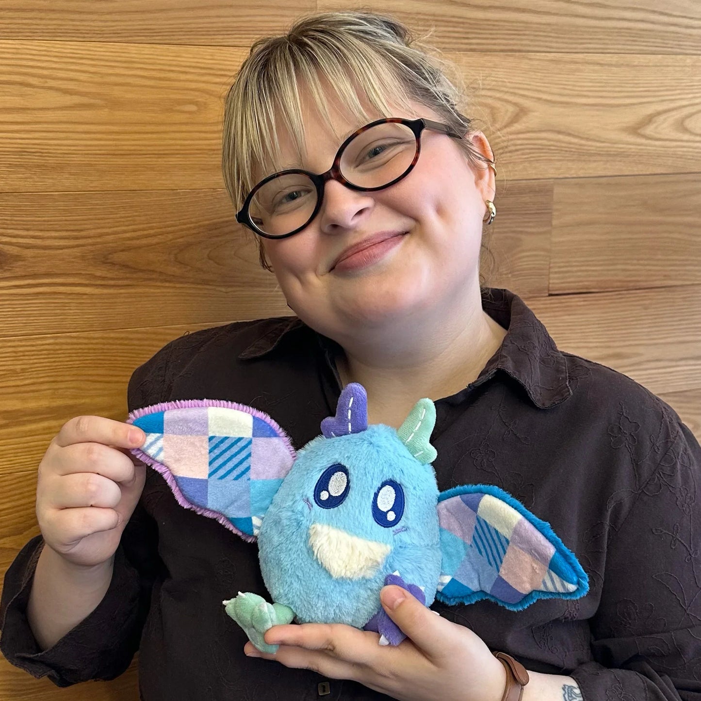 A person with glasses and a dark brown shirt holds a Squishable Alter Egos Series 21 Quilted Mothman plush with sky blue fur, mismatched antennae, and patchwork quilted wings in front of a wooden wall.