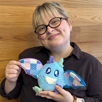 A person with glasses and a dark brown shirt holds a Squishable Alter Egos Series 21 Quilted Mothman plush with sky blue fur, mismatched antennae, and patchwork quilted wings in front of a wooden wall.