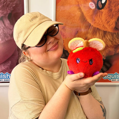 A person wearing a beige cap and glasses holds Squishable Snacker Fruit Fly S1E3 limited edition plush shaped like a red apple with citrus slice wings and purple feet. in front of a colorful plush bear poster.