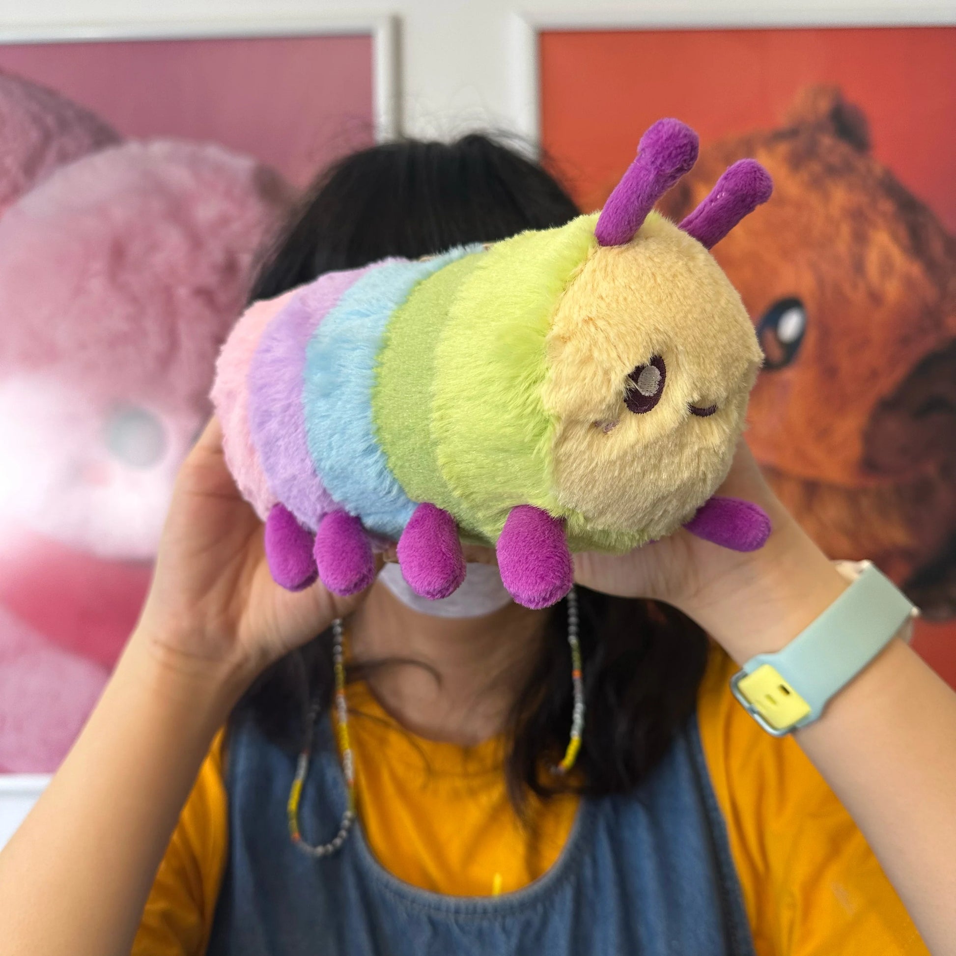 A person holding a Squishable Snacker Rainbow Caterpillar S1E5 Limited Edition plush with pastel rainbow segments, magenta antennae, and small legs.