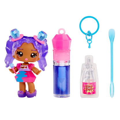Yummiland x Ring Pop lip gloss doll with purple hair featuring gemstone accents, wearing a pink dress, shown with DIY lip gloss kit components including blue lip gloss tube, scented liquid packet, stirring spoon, and keychain ring.